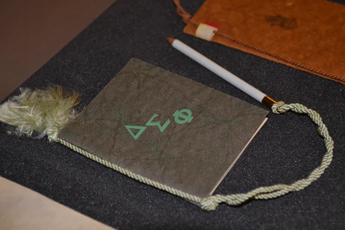 close up of gray dance card with three Greek symbols on the cover with an attached white pencil and tassel. 