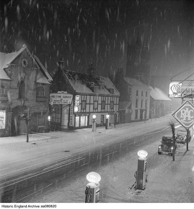A black and white photograph of a snowy street at night. Snow is falling and the roofs, pavements and edges of the road are covered in a dusting of snow. The road stretches from left to right diagonally across the photograph. In the foreground are two petrol pumps with 'Esso' illuminated on top of each. To the right, signs including an RAC sign hang over the pavement from a building which is out of shot. Beyond this are two parked cars. On the opposite side of the road is a row of buildings. In the centre is a timber framed building. To the left is a sign for Melton Garage, and to the right is a church tower which is just visible against the dark night sky.