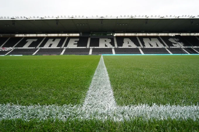 View of Pride Park Stadium's well-maintained football pitch,  with "THE RAMS" spelled out on the black and white stadium seats in the background.