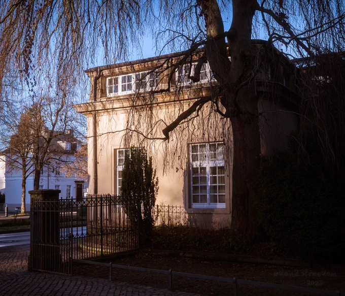 The serene and nostalgic atmosphere of a classical building at the entrance to picturesque gardens, emphasizing the interplay of warm sunlight and cold winter tones. The composition highlights architectural elegance framed by natural elements, evoking a sense of calm and timeless beauty.
—
Light guards the threshold,  
  Songs echo through frozen air.