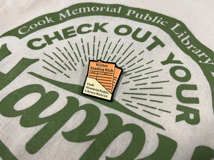 An image of an orange and yellow enamel pin that looks like an old-timey library catalog and says "Winter Reading Club and Cook Memorial Public Library District" on top of a tote bag that reads "Cook Memorial Public Library -- Check Out Your Happy"