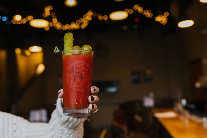 A person wearing a knitted sweater holds a glass of Bloody Mary at a bar decorated with twinkling lights.