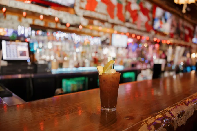 A Bloody Mary garnished with lemon sits on a wooden bar counter, with a blurred background of a vibrant bar setting featuring colorful decorations and a well-lit display of various bottles.