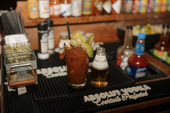 A Bloody Mary cocktail garnished with olives and celery sits on an "ABSOLUT VODKA Cocktail Perfection" bar mat surrounded by condiments and bar tools.