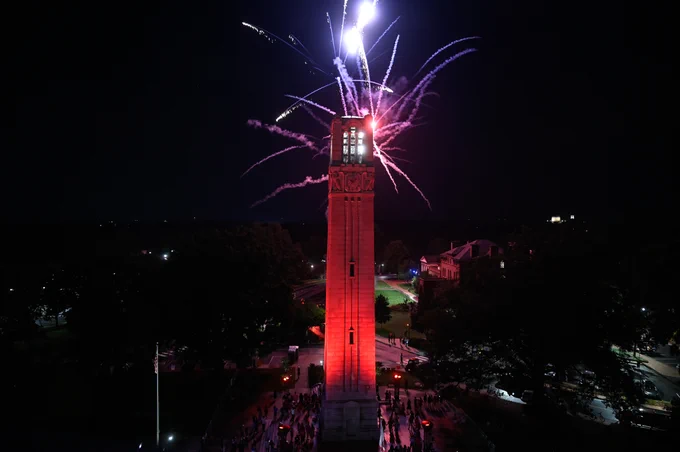 Tall clock tower illuminated in red with colorful fireworks bursting behind it against a dark night sky.