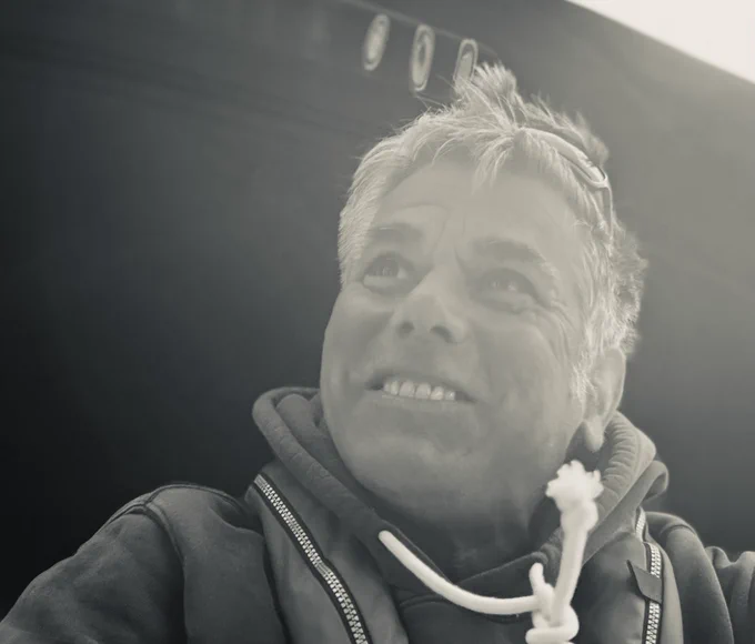 Portrait eines Mannes in schwarz-weiß. Der Mann hat leicht, ganz leicht graue Haare lächelt in sich gekehrt zufrieden, leicht debil und blickt auf die linke obere Bildecke. Er trägt einen Hoodie und eine automatische Rettungsweste. Der Hintergrund ist grau, das Bild ist von schräg unten aufgenommen.