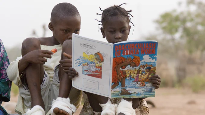 The image shows two children reading a book together about Guinea worm disease.