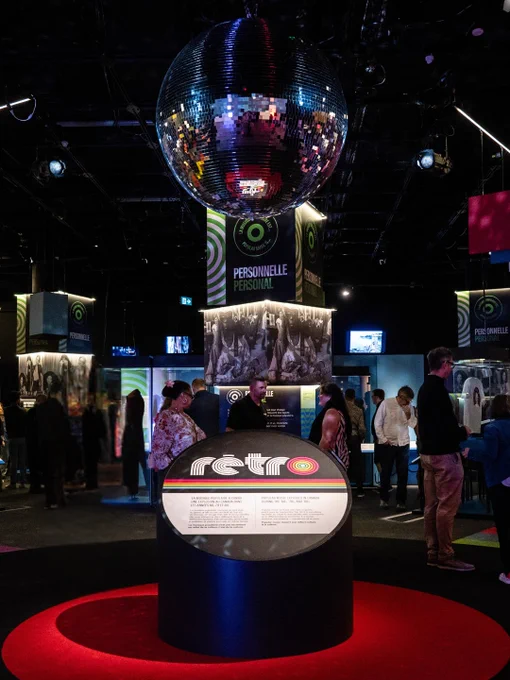 À l’intérieur d’une salle d’exposition du Musée, les visiteurs interagissent avec diverses présentations sous une grande boule disco. Au premier plan, un podium d’information arbore de l’information au sujet de l’exposition Rétro.