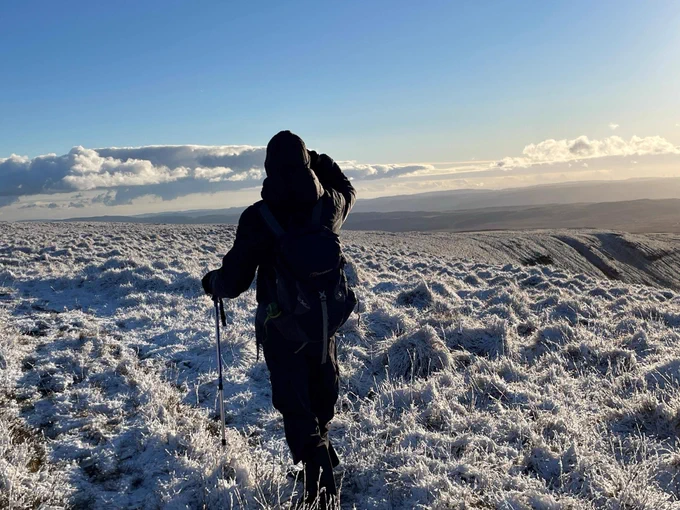 A person hiking over a frost-covered hilly landscape with a backpack and trekking pole, under a mostly clear blue sky.