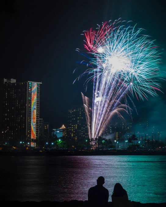 Two people sitting by the water, watching a vibrant fireworks display over a city skyline at night.