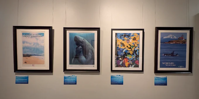 Four framed ocean-themed artworks hang on a gallery wall, each with a small blue placard beneath.