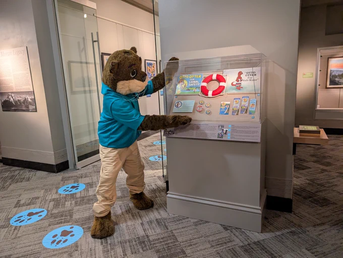 A costumed otter interacts with an educational display about Otto Otter in a museum gallery.