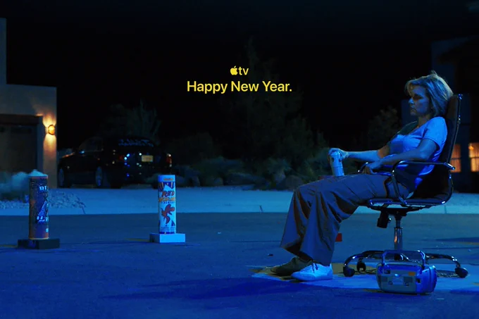 From Pluribus. Rhea Seehorn as Carol slouches on a chair outside on the street with a can of beer in her hand. She is facing two fireworks propped on the street, ready to be launched into the sky. Text on the top of the image reads, "Happy New Year" underneath the Apple TV logo.