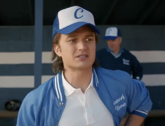 Steve Harrington stands on a baseball field with a blue hat and jacket that reads “Coach Steve”