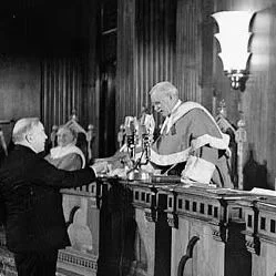 Black and white photograph showing four men in a wood-paneled chamber with tall carved columns and a wall light fixture.

One man in a dark robe stands on the left at a wooden dispatch box, holding a paper in his right hand and raising his left hand.

Across the table from him sits a man in black robes with gold embroidery and a tricorn hat on the red Woolsack.

Two additional men in robes and wigs sit on raised ornate wooden benches behind the man on the Woolsack, one holding papers.

The image is sharply focused on the standing man and the seated central figure.