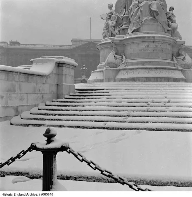 A black and white photograph of snow covering steps leading up to a large monument. The monument has sculptures on top of a base with the inscription 'Victoria Regina Imperatrix'. The words 'Happy New Year' is written in snow around the base of the monument. In the foreground is a post and chain fence.