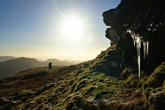 A person walking on a grassy hill at sunrise, with a rocky outcrop and icicles to the side, overlooking a distant mountain range.