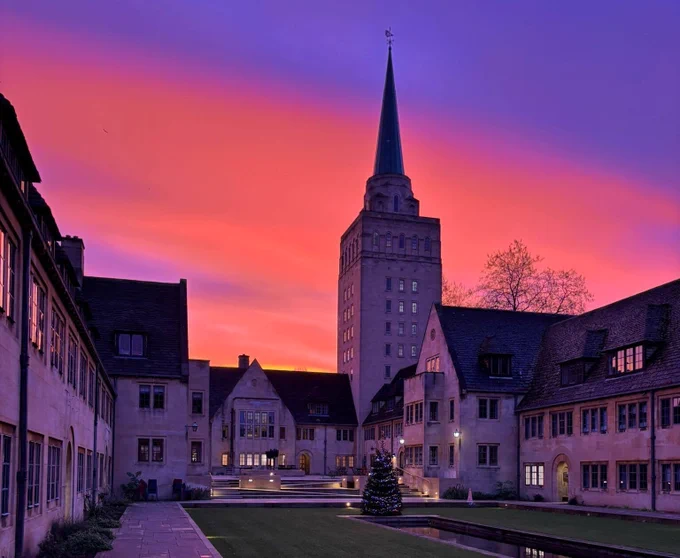 A rosy sunrise over Nuffield College, with the College tower against an orange, pink and purple sky. Some lights are on in the building, and the windows reflect the sunrise.