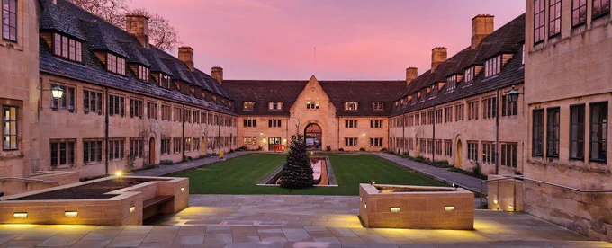 A warm pink sunrise over the lower quad of Nuffield College. Lights are still lit and a Christmas tree sits in front of the pond. The main gate and the new Porters' Lodge is in the background.
