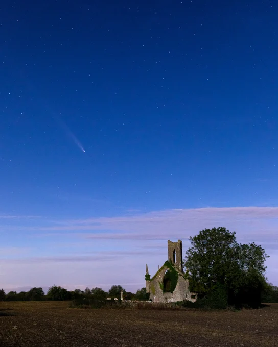 A comet with a bright tail streaks across a starry night sky over a field. Below, the ruins of an old stone church, partly covered in ivy, stand beside a large tree. The scene is illuminated by a soft glow, creating a serene and mystical atmosphere.