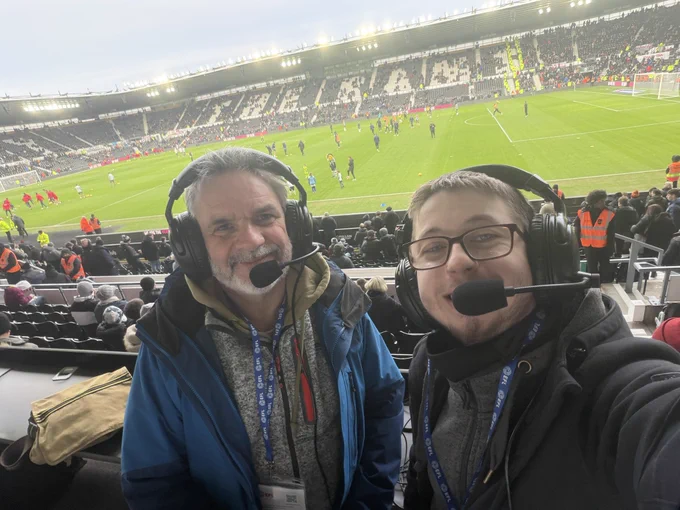 A landscape of your commentary team Matty and Stuart with the pitch in the background with fans scattered around the east stand at the very back