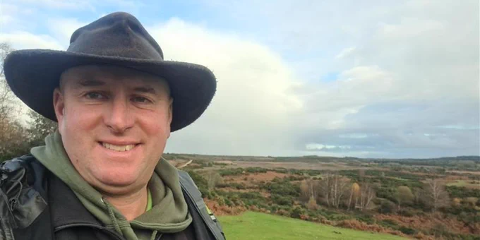 New Forest NPA ranger smiling at the camera, wearing a hat and outdoor clothing, with a scenic, open landscape in the background.