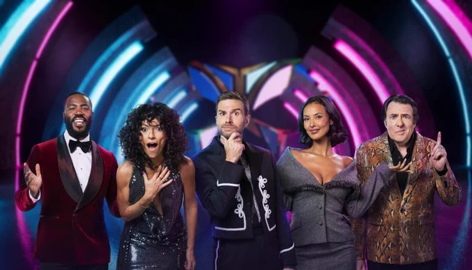 Image of The Masked Singer panel in front of a blue and pink studio light - from left to right - Mo Gilligan (wearing a burgundy velvet jacket, white shirt and bow tie), Davina McCall (wearing a silver dress and black curly wig), host Joel Dommett (wearing a black and white matador-type suit), Maya Jama (wearing a grey off the shoulder suit) and Jonathan Ross (wearing a brown snake-skin jacket and black shirt)