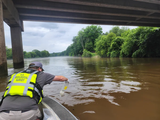 Man collecting a water sample from inside of a small boat, under a bridge.