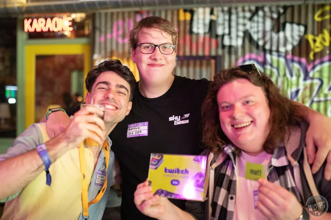 Three people smiling at a music and creator event, posing together in a colourful venue. One holds a Twitch Creator Club card while another raises a drink. Bright lighting, neon signs, and street-art décor create a club-style atmosphere.