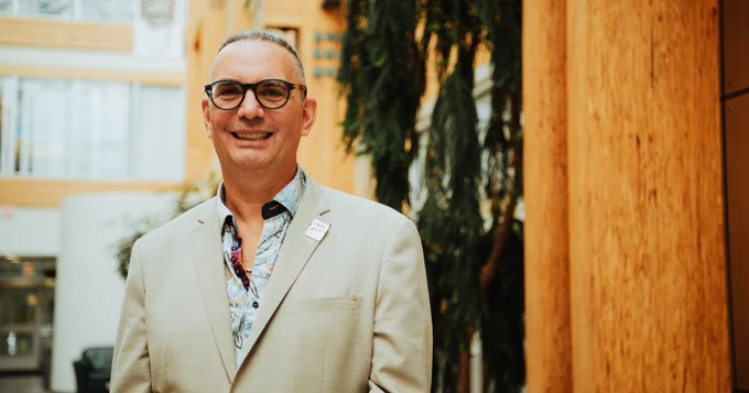 Individual wearing glasses and a beige blazer standing in a sunny atrium with green plants and wooden decor, smiling at the camera.
