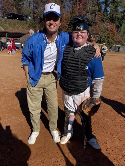 Joe Keery and Jake Connelly on a baseball field. Keery is wearing a blue jacket, white shirt, khaki pants, white shoes and a blue and white baseball cap. Connelly is wearing catcher's gear, a blue baseball jersey and white pinstriped pants.