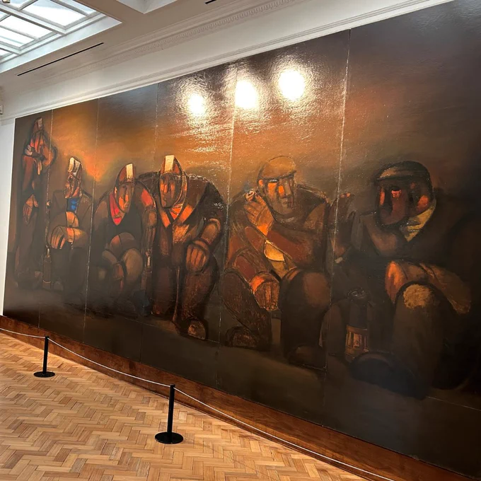 On the wall of a gallery is a large painting in earthy brown tones which is made up of 6 panels. Altogether, the painting is almost 3 metres tall and over 6 metres wide. The painting shows 6 miners: all but one crouch down while the miner furthest to the left stands up. 