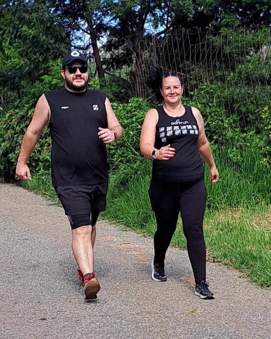 Two parkrunners wearing black best jogging on a pathway with lush greenery in the background. 