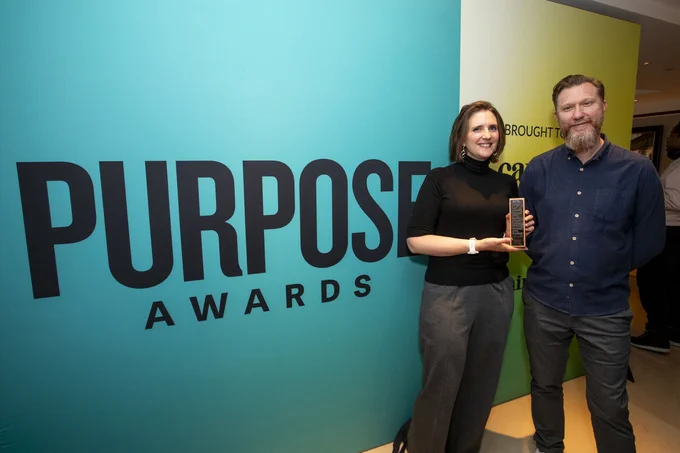 Two individuals smiling and holding an award at the Purpose Awards event, standing in front of a bright blue background with the event's name prominently displayed.