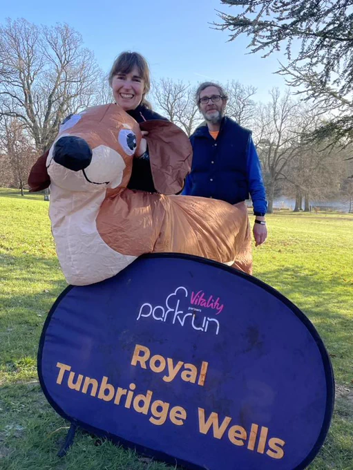 Two parkrunners, one in an inflatable meerkat costume, smiling beside the Royal Tunbridge Wells parkrun's purple panel on a sunny day with grass and trees in the background. 