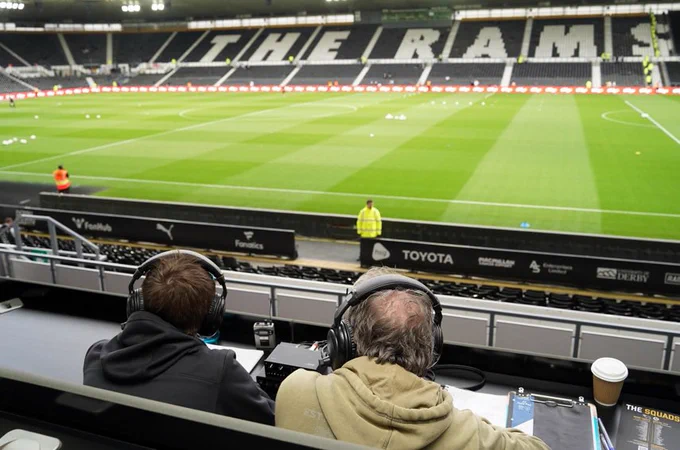 Landscape photo of 2 members of the DCFCAD team doing some pre match testing and talking.

In the background is the luscious green pitch of Pride Park and then behind that is the East stand with the patterned seats reading THE RAMS being fully visible