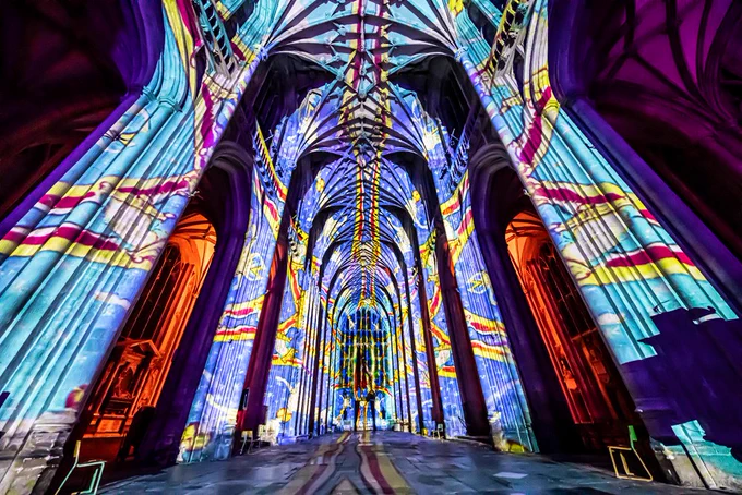 Vibrant light projections illuminate a cathedral's interior, featuring swirling patterns in purple, blue, and orange, creating a mesmerising, ethereal atmosphere.