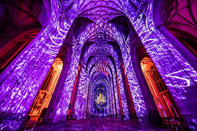 A cathedral interior illuminated with vibrant purple and orange lights, casting intricate patterns across arched ceilings and walls, creating a mystical ambience.