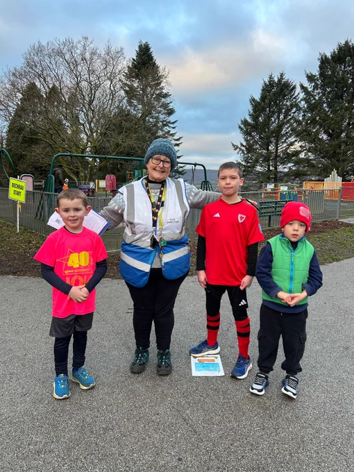Three junior parkrunners taking pictures with a run director, standing on a path with playground equipment in the background.