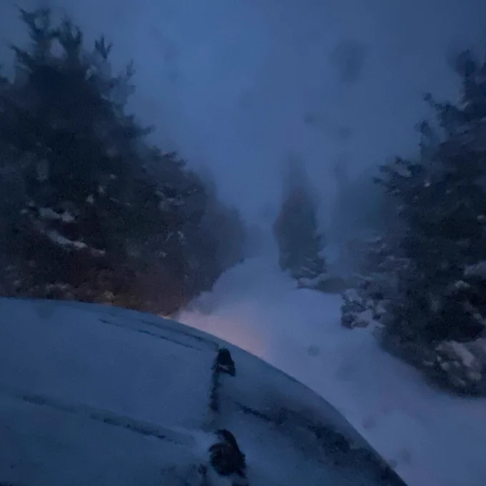 Snow-covered landscape during twilight with visible car partially covered by snow.