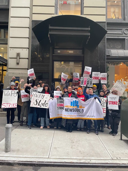 A few dozen people holding a News Guild of New York banner and strike signs. Some display fists of solidarity.
