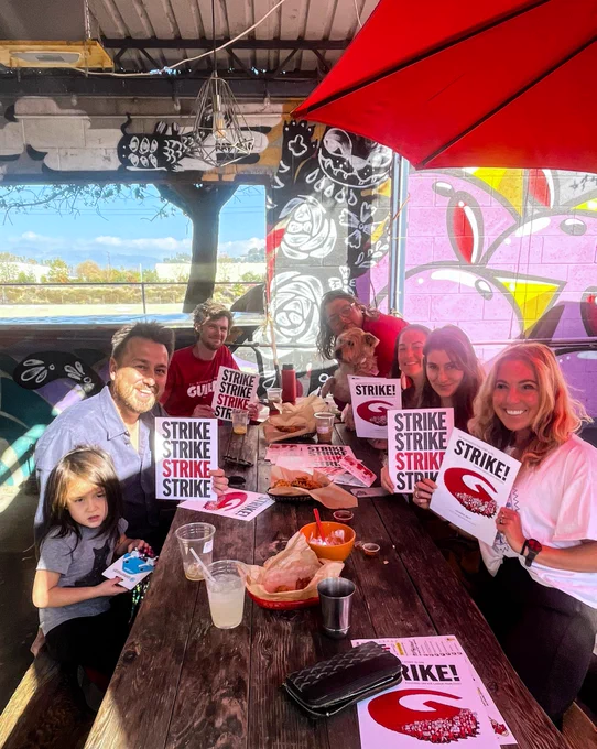 A group sitting at a table holding strike signs and posters with baskets of food.
