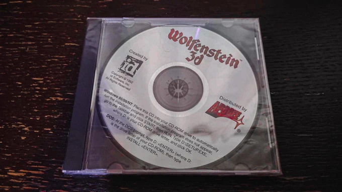 
This image displays a CD-ROM of the classic first-person shooter game "Wolfenstein 3D," resting inside a clear jewel case. The disc is prominently labeled with the game's title in a bold, stylized font. Logos for the creators, id Software, and the distributor, Apogee, are also visible. Instructions for Windows 95/98/NT users and DOS users are provided, detailing how to install the game from the CD-ROM. The CD itself has a simple yet iconic design, reflective of the early era of PC gaming. The dark textured surface on which the CD case lies adds a contrast that highlights the vintage appeal of this gaming artifact.