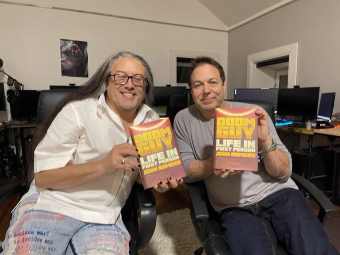 
In the image, video game developers John Romero and Scott Miller are seated next to each other, each holding a copy of John Romero's book titled "DOOM GUY: Life in First Person." 