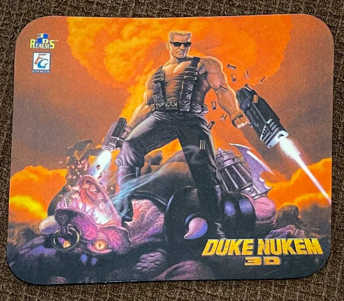 The image features a mouse pad with the iconic artwork from the game "Duke Nukem 3D."