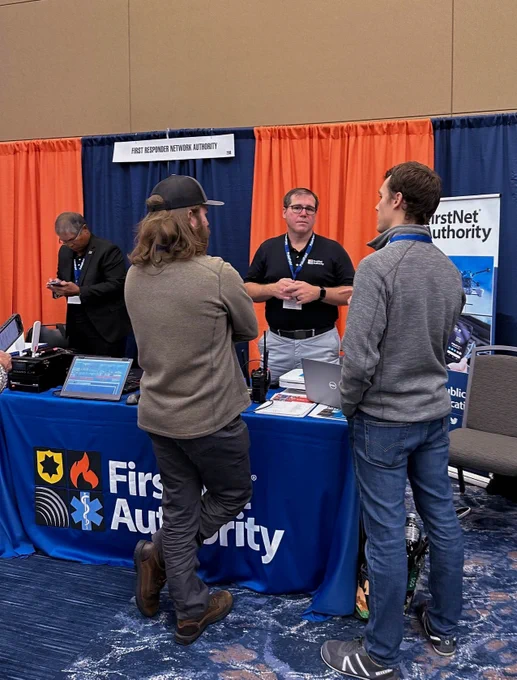 A FirstNet Authority representative behind a booth, with the FirstNet Authority logo on it, speaking to two participants at the IAFC TSI Conference.