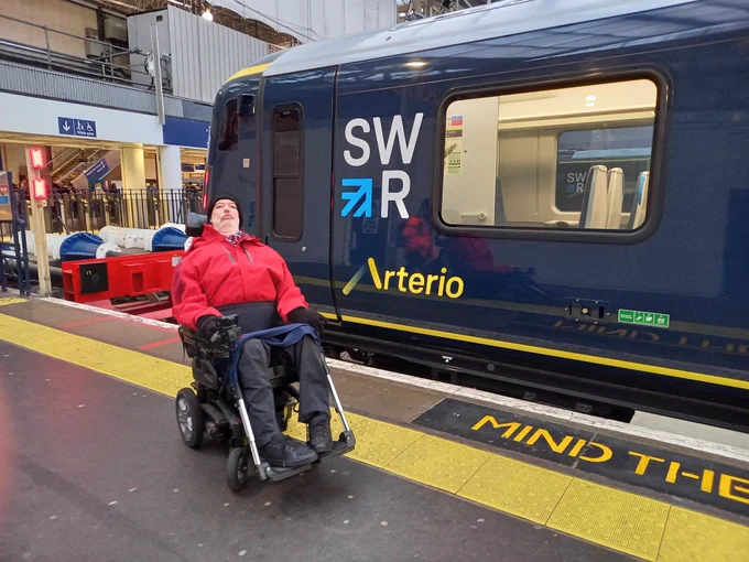 Alan in his wheelchair, in front of a new train. It is dark blue with the white SWR logo and the name "Arterio" in yellow on the side. It is next to a platform edge. Alan is wearing a red coat and dark trousers. 