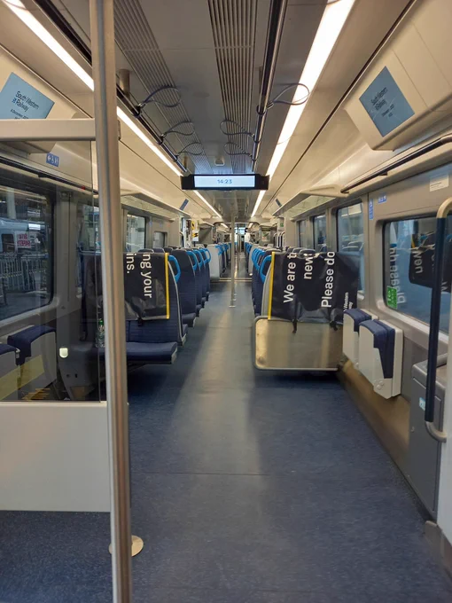 A view down the aisle of the new train. It has cream coloured walls and ceiling, grey floor, silver poles and dark seats with blue grips on them. The wheelchair space is large with room to move around. 