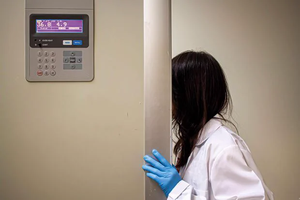 A female scientist wearing a white laboratory coat and blue nitrile gloves opens the door of an incubator.