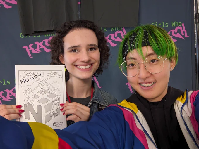 Bia and Mara standing together, with Mars NumPy comic held up, in front of the Girls Who Chose booth.